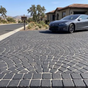 Ground-level view of a wide residential driveway featuring antique-finish dark stone cobblestones laid in an overlapping fan pattern, with a dark grey sedan parked in front of a stucco home.