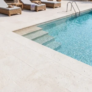 Natural white limestone pavers surrounding a pool with built-in steps, rattan lounge chairs, and Mediterranean-style architecture at a U.S. home