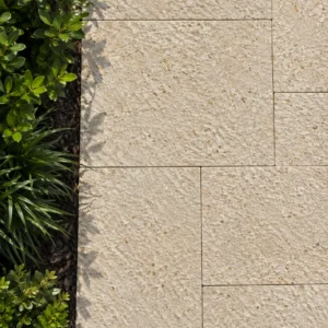 Direct overhead close-up of warm beige, deeply textured paving stones meeting a dark soil bed filled with small green plants, highlighted by a leafy shadow.