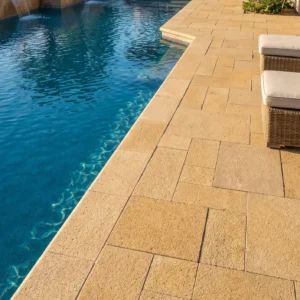 Low-angle view along the edge of a bright blue swimming pool bordered by golden-tan stone pavers featuring a rough texture laid in a multi-size ashlar pattern.