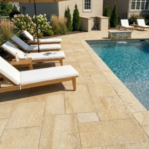 Eye-level view of a residential pool deck paved with textured golden yellow limestone in an ashlar pattern, featuring wooden sun loungers with white cushions next to a swimming pool.