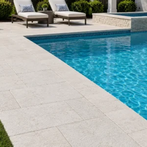 Angled view of a bright white limestone pool deck and straight coping edge bordering a bright blue swimming pool, featuring a raised spa with a waterfall and two white lounge chairs.