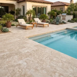 Mid-level shot across a bright blue swimming pool bordered by oversized, textured beige stone slabs exhibiting distinct fossil patterns, leading to twin wooden lounge chairs, tropical landscaping, and a Mediterranean villa.