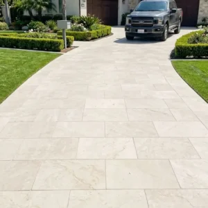 Vertical orientation showcasing a wide, curving residential driveway paved with smooth, light-colored rectangular stone blocks, leading past manicured boxwood hedges and palm trees toward a Mediterranean-style home where a dark pickup truck is parked.