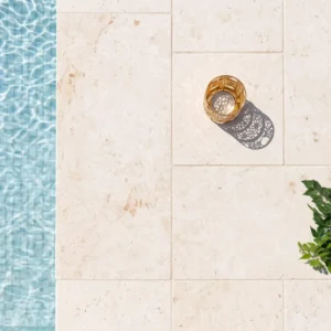 Cream stone pool deck pavers in a French pattern beside rippling pool water, with a gold lantern and green leaves