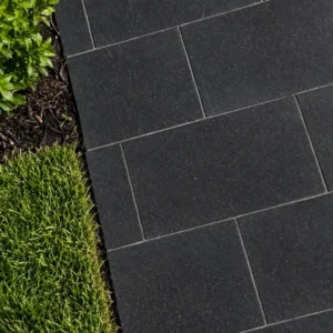 Modular rectangular black stone pavers with fine light flecks, bordered by fresh green grass and a mulched shrub bed