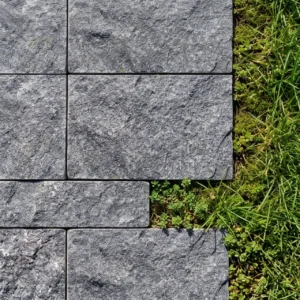 Direct overhead view of highly textured dark grey stone blocks arranged in an offset pattern, meeting a bright green lawn with a slightly jagged boundary.