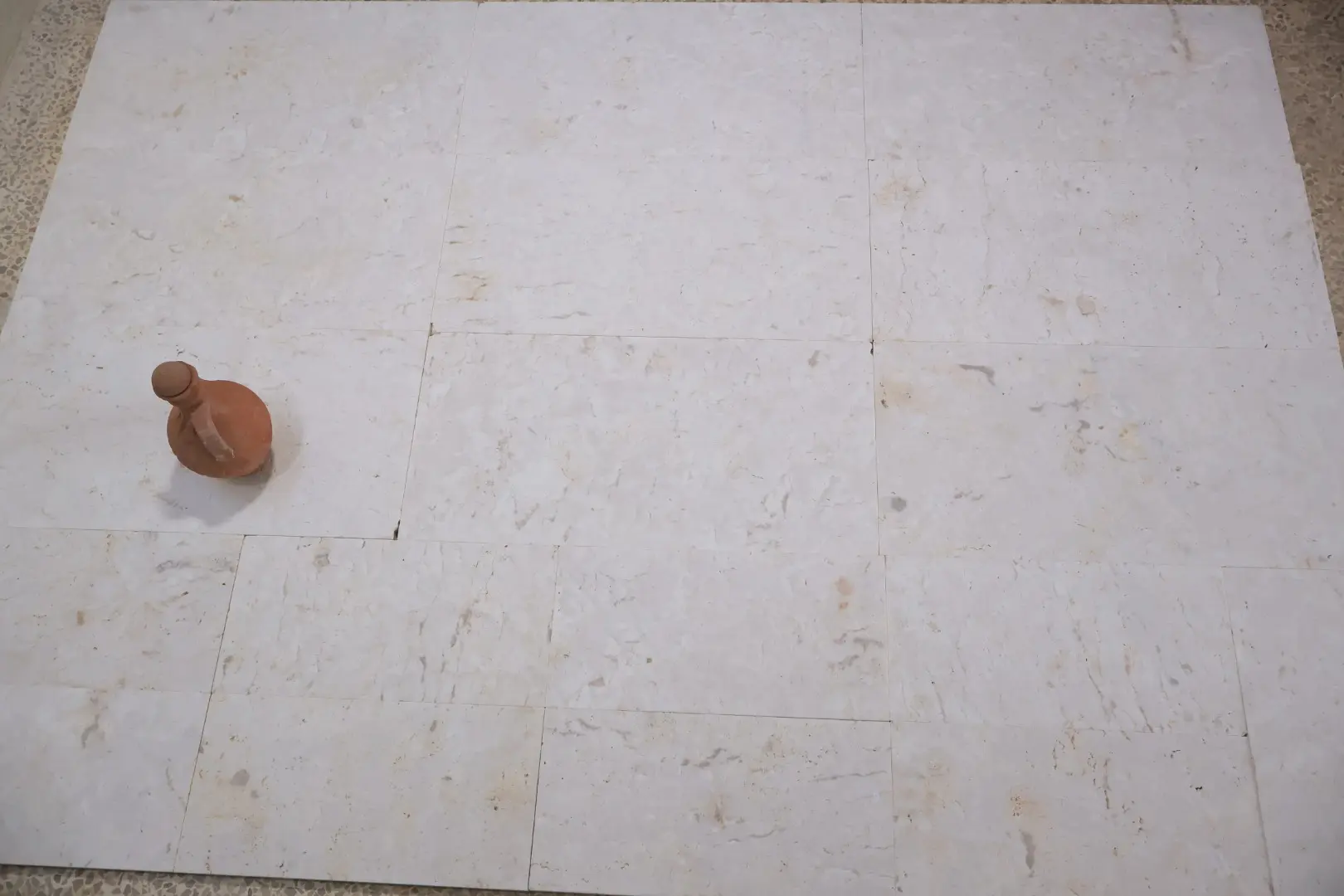 A small terra cotta jug rests on a light-colored marble tile floor.