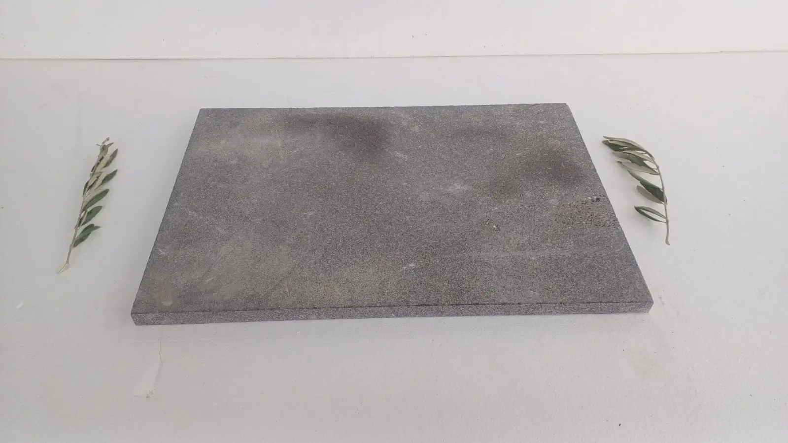 A dark grey speckled stone slab rests on a white surface with olive branches on either side.