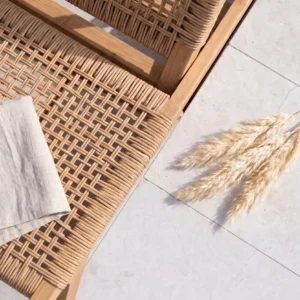 Top-down close-up of a woven wooden patio chair and dried pampas grass resting on smooth, bright white limestone pavers in harsh sunlight.