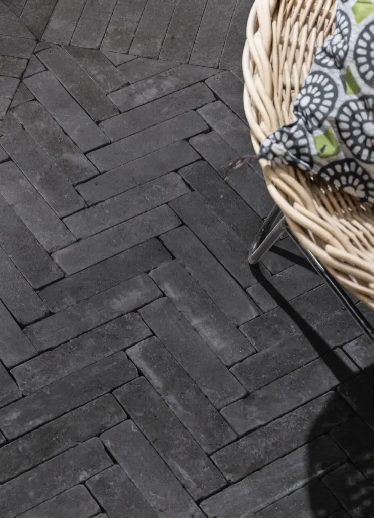 Black Basalt Plank Setts – Brushed (8″ × 2″ × 2″) 1 Brushed black basalt setts laid in a herringbone patio pattern beside a rattan chair