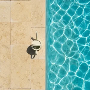 Top-down detail view of a bright turquoise swimming pool edge meeting warm golden Ambra Reale brushed 24-grit stone pavers, with a modern watering can resting on the coping.