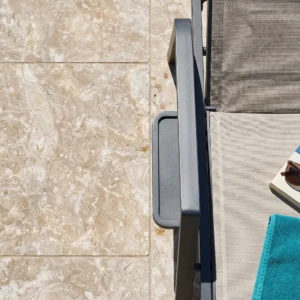 Direct overhead top-down close-up detail of Ocean Reef Shellstone pavers with a brushed 60-grit finish, fossilized shell inclusions, and beige-cream tones, alongside a dark metal pool lounger.