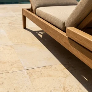Close-up ground-level view of a teak lounge chair with beige cushions resting on square stone pavers featuring a warm golden-amber base, delicate dark veining, and a tactile brushed 24-grit finish.