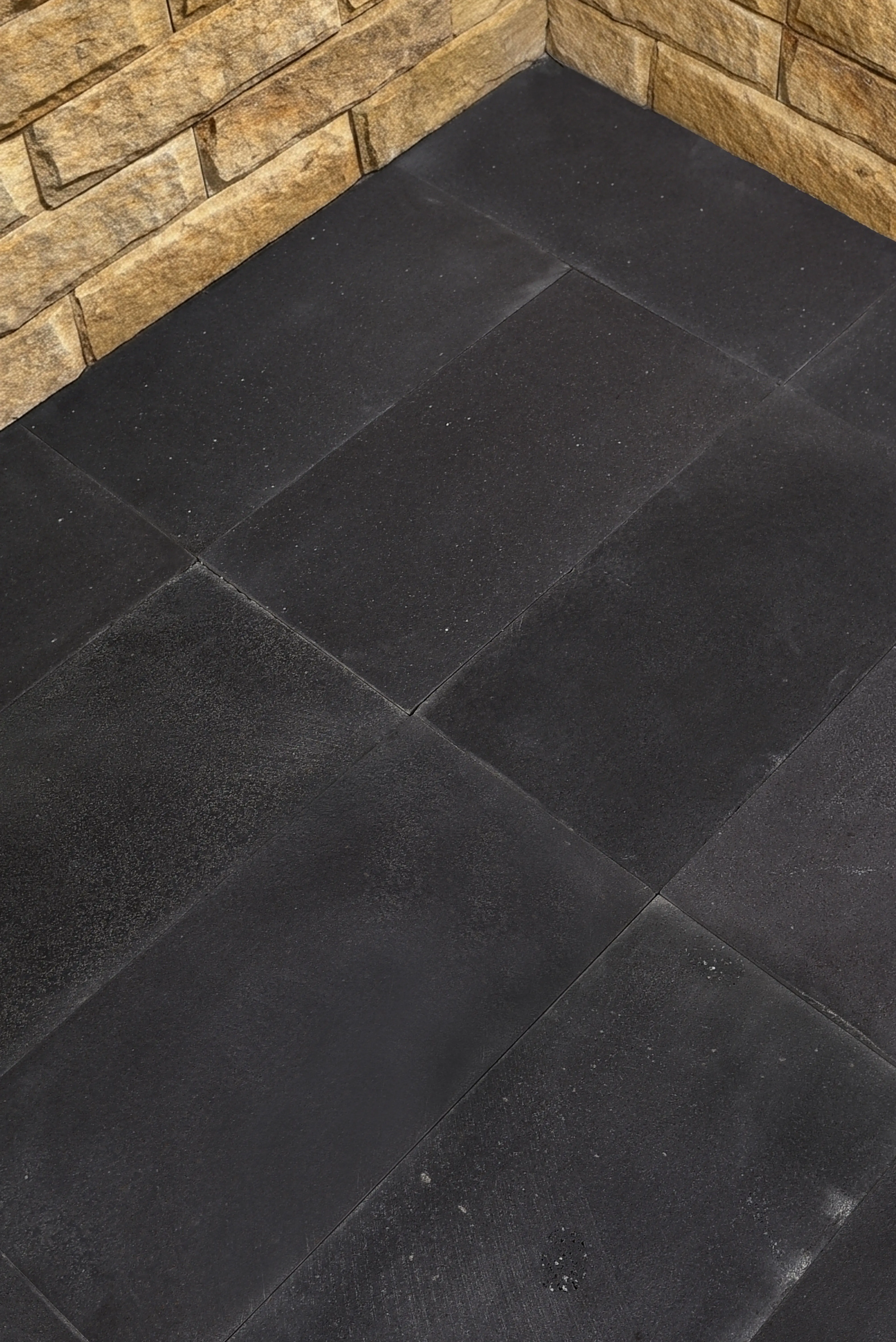 Close-up of interlocking black rubber flooring tiles next to a textured stone wall, a reference for blue black slate paving slabs.