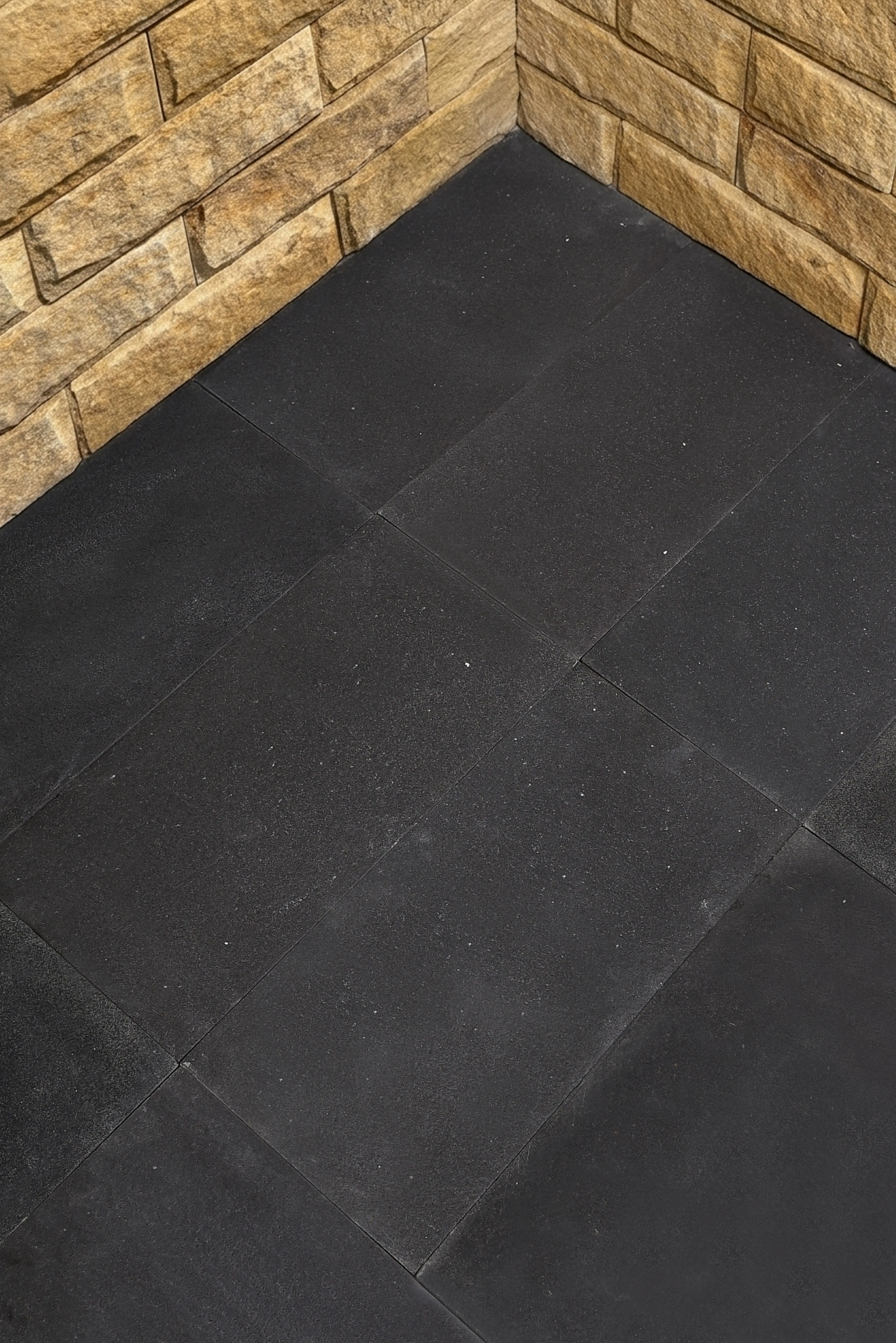 Dark square floor tiles meet a tan stacked stone wall at a corner, an example for lagos blue limestone.