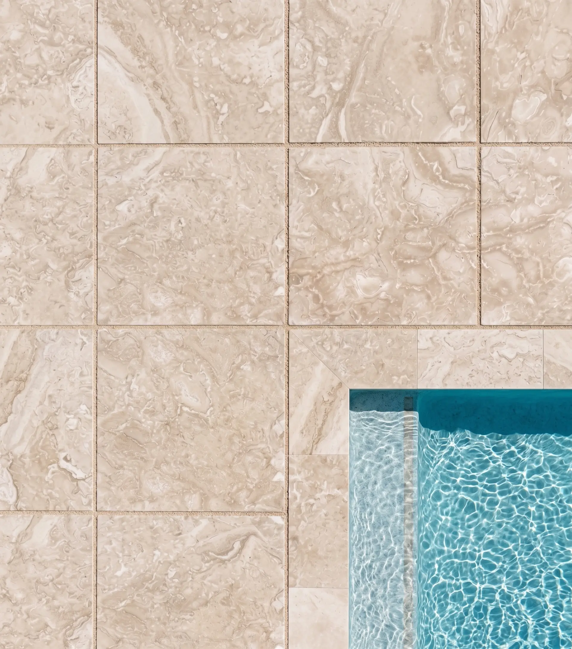 Light travertine tiles bordering clear blue pool water with sunlit ripples, a reference for cleaning travertine pavers.