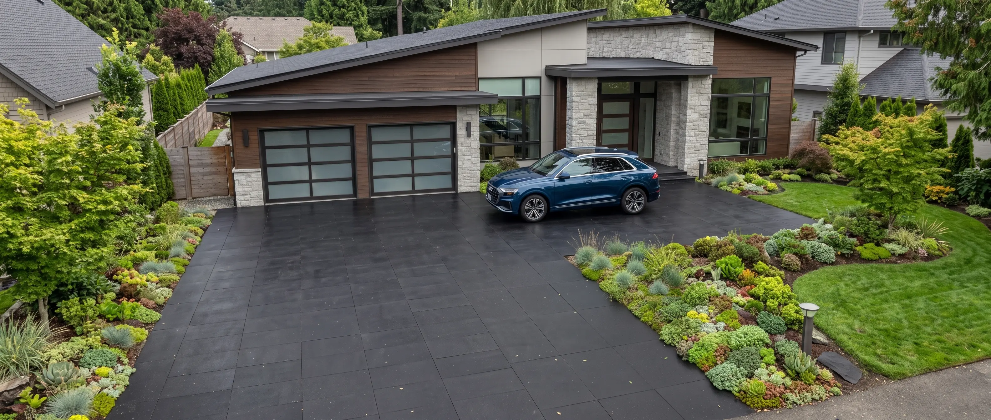 A modern home's dark paver driveway with a blue SUV and abundant, textured plantings.