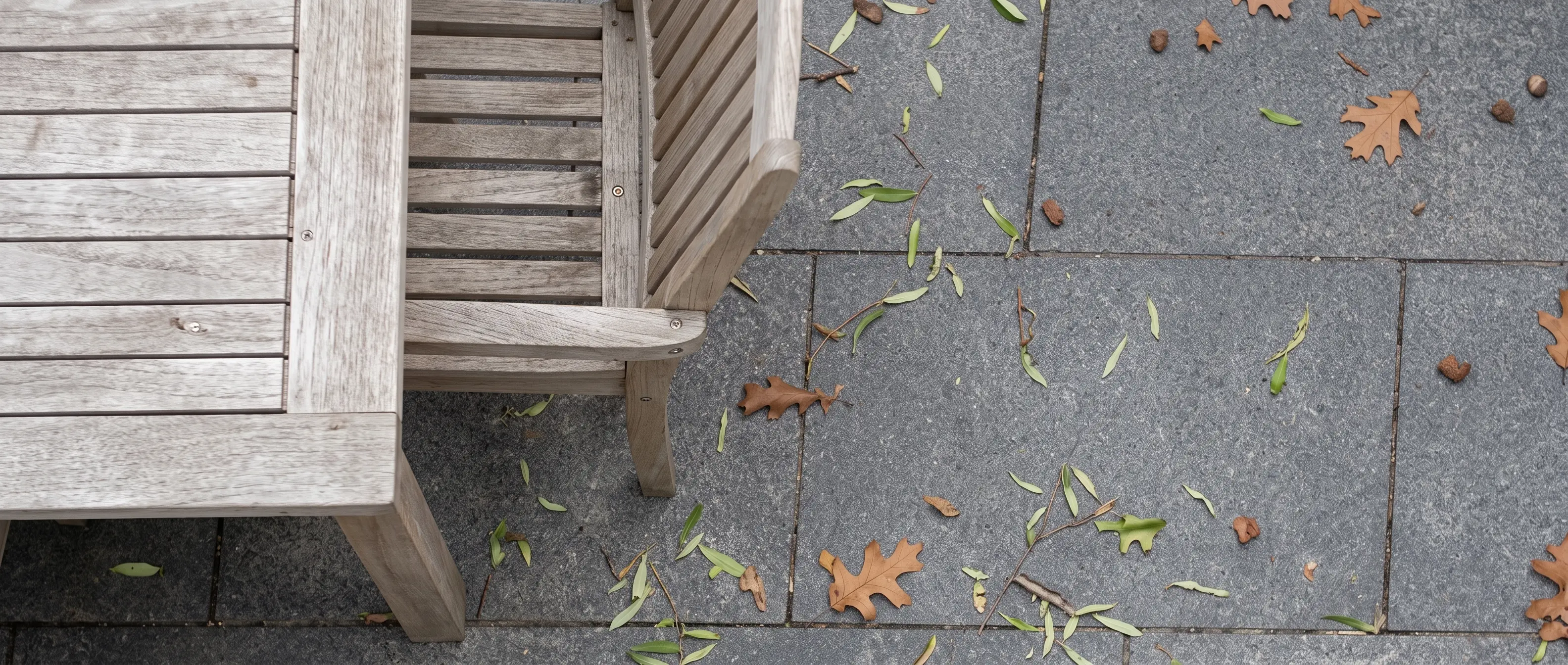 Wooden outdoor bench placed on a dark stone paved patio with fallen leaves.