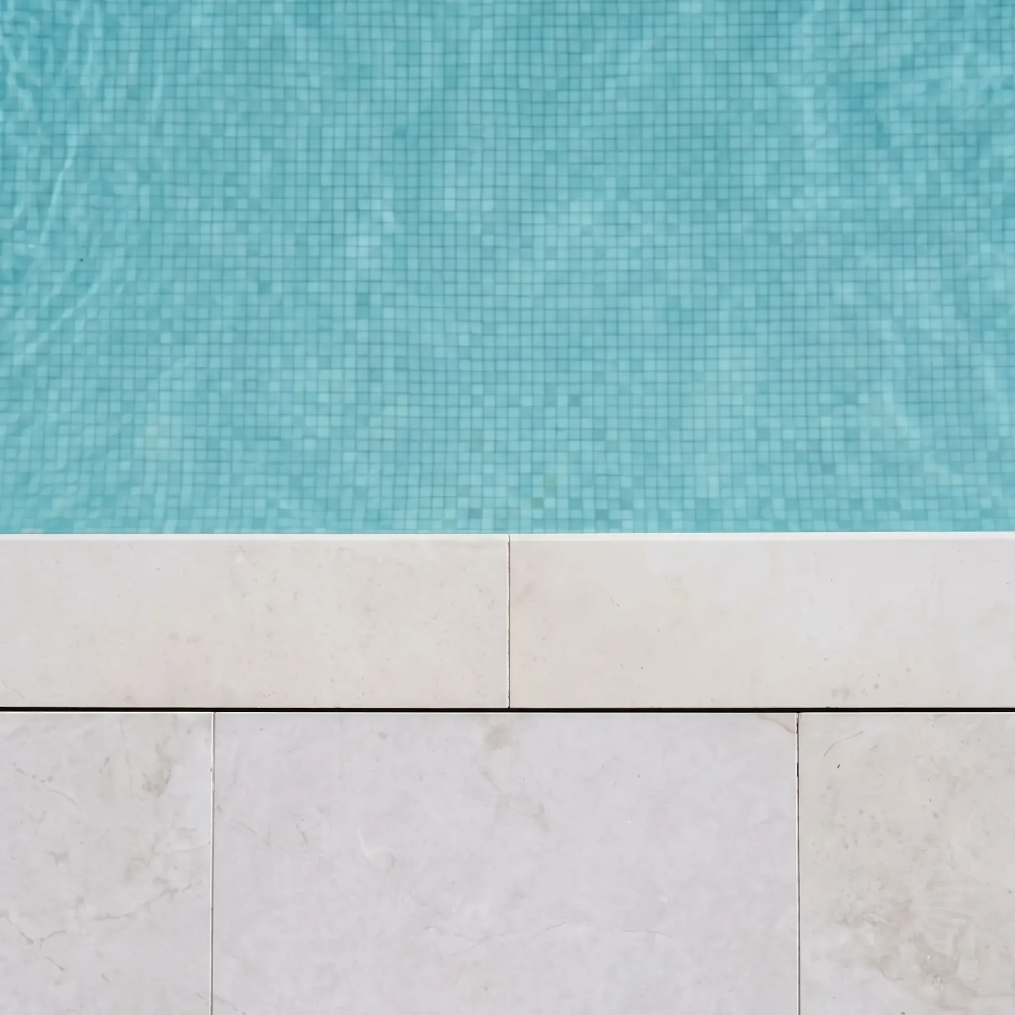 top-down pool edge: cream limestone-look coping and square pavers against aqua mosaic-tiled pool water