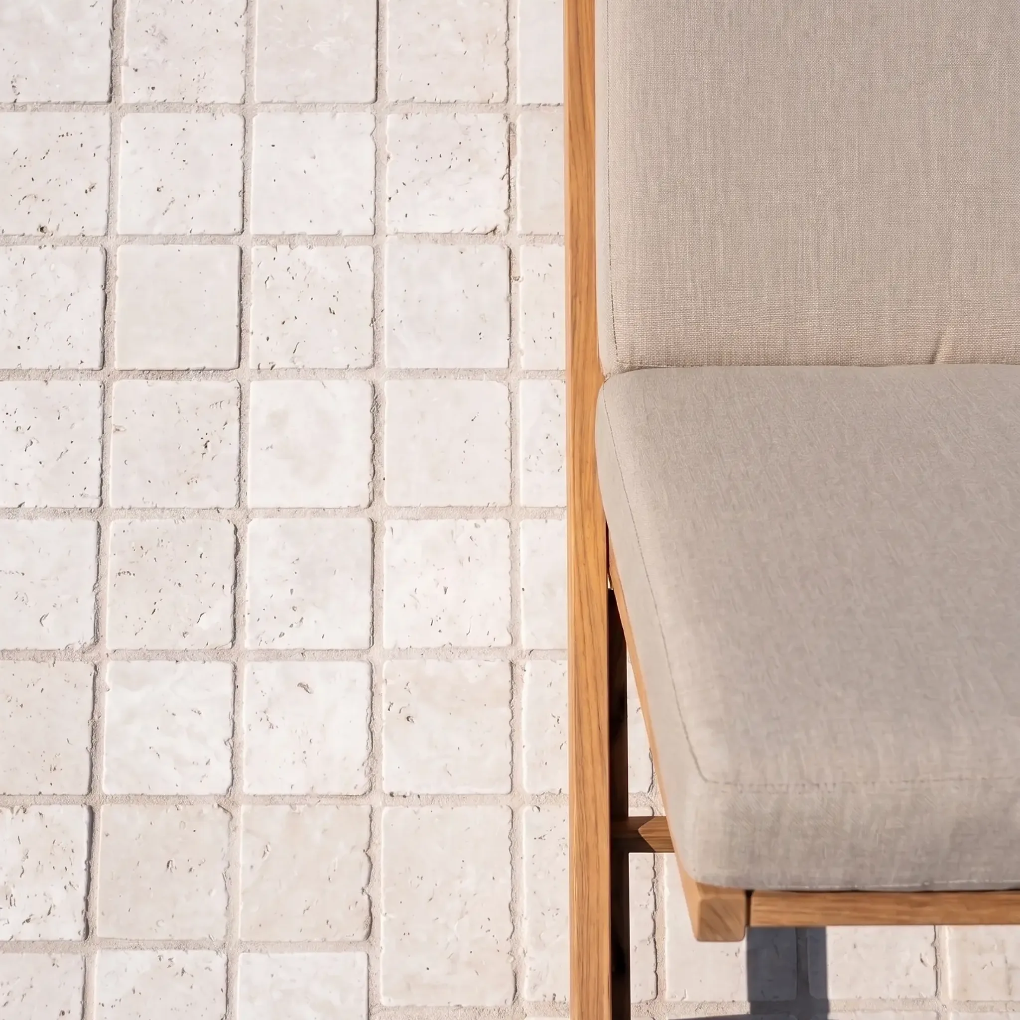 Close detail of cream pitted limestone tumbled cobbles with sand joints beside the corner of a teak lounger with beige…