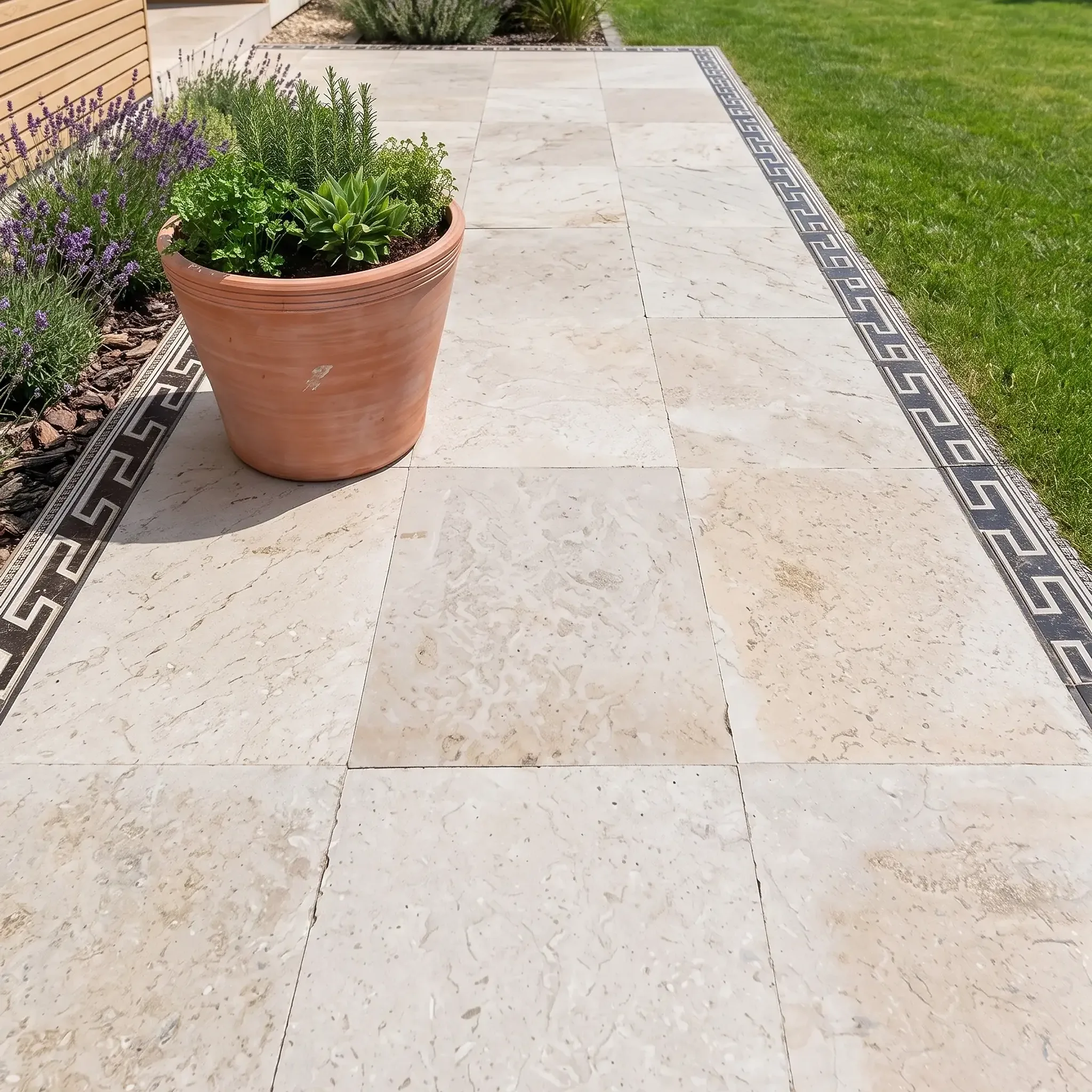 Light beige travertine paver walkway with black Greek key mosaic border, terracotta herb pot, lavender and lawn beside a…