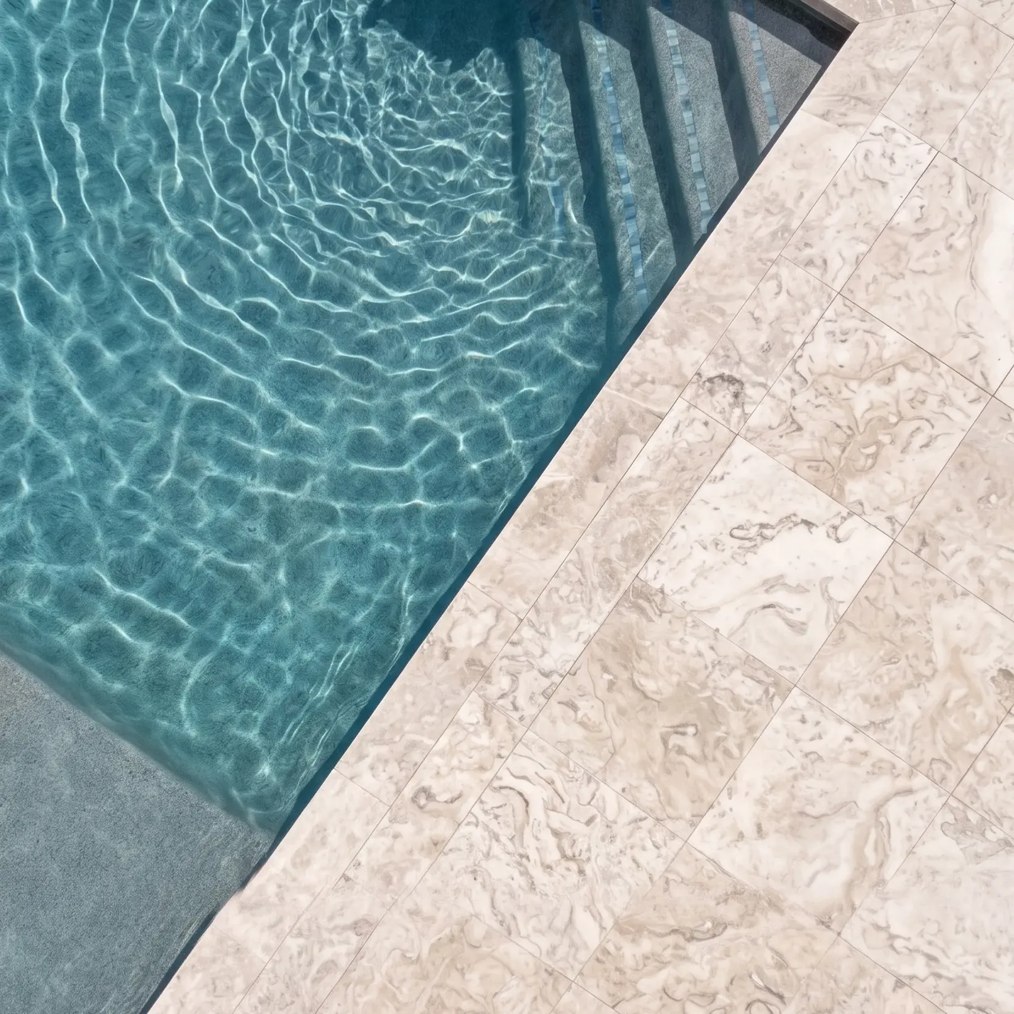 Overhead view of pool corner with steel ladder, rippling water and beige fossil-marked limestone pavers forming deck and…