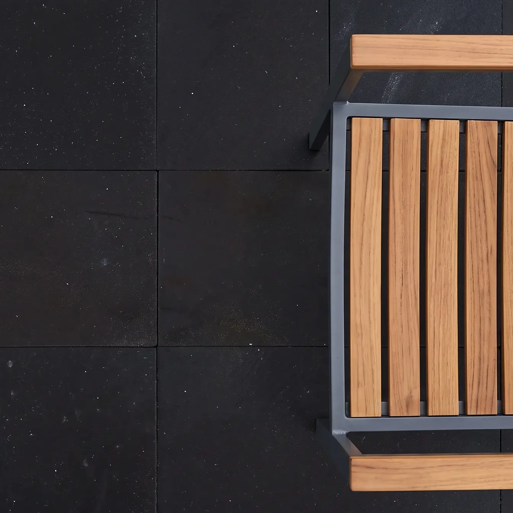 Top-down view of black stone pavers with white speckle and a teak-and-aluminium patio chair; even lighting.