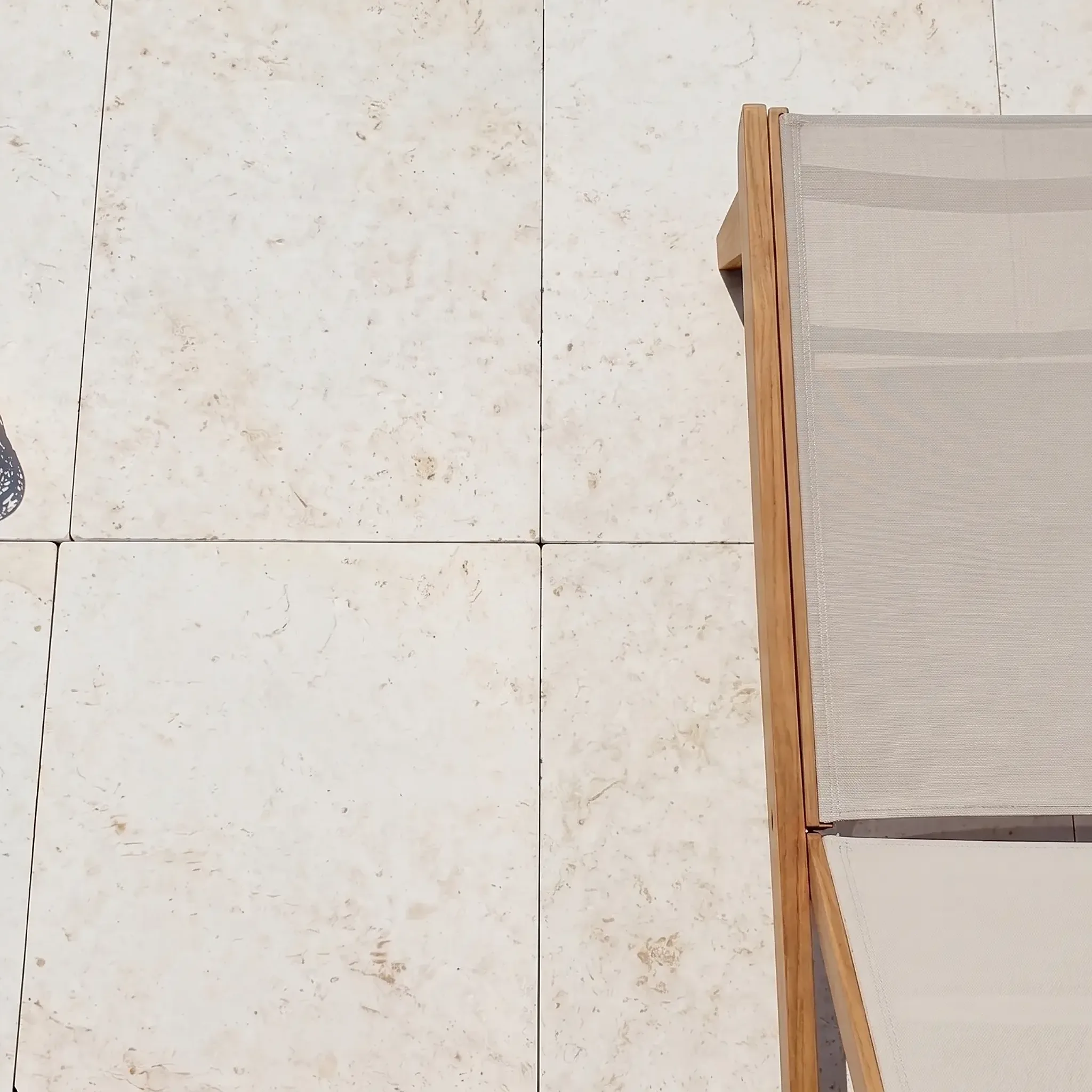 Overhead photo of white limestone pavers with pitted texture, a teak sling sunbed, gold lantern and a leafy branch on a…