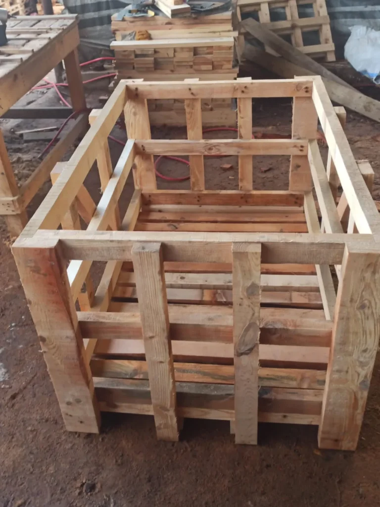 Empty deep wooden crate frame with slatted sides in a workshop, with stacked timber behind.