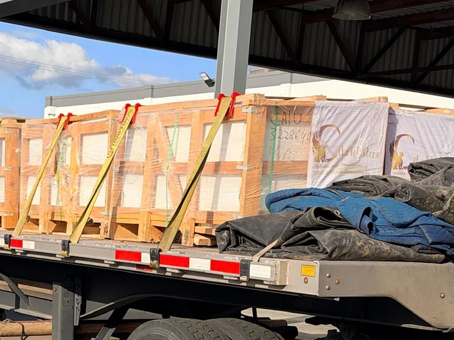 Film-wrapped wooden crates held down with yellow straps on a flatbed trailer, one crate hand-marked CAIU 322864 0.