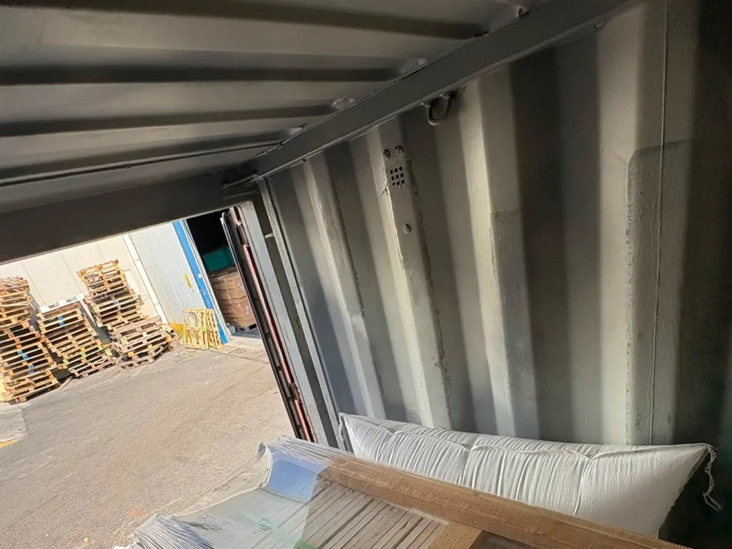 Film-wrapped crates near a container door, with a white air bag against the wall and a yard visible through the door.