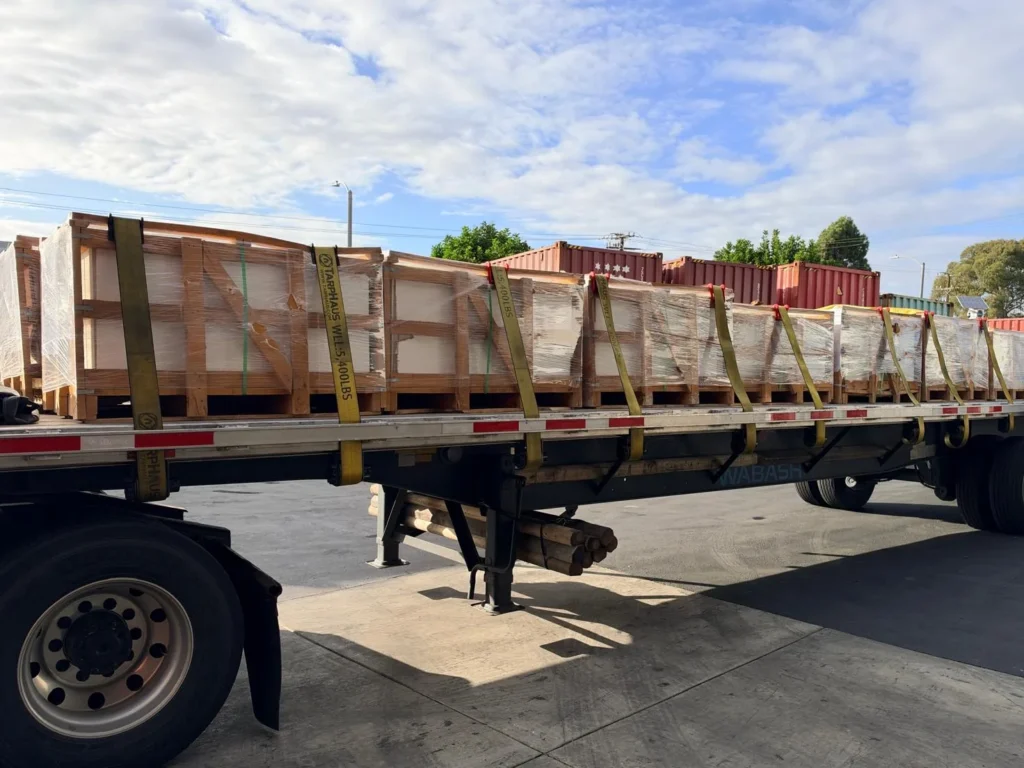 Flatbed trailer loaded with film-wrapped wooden crates, held down with yellow straps.