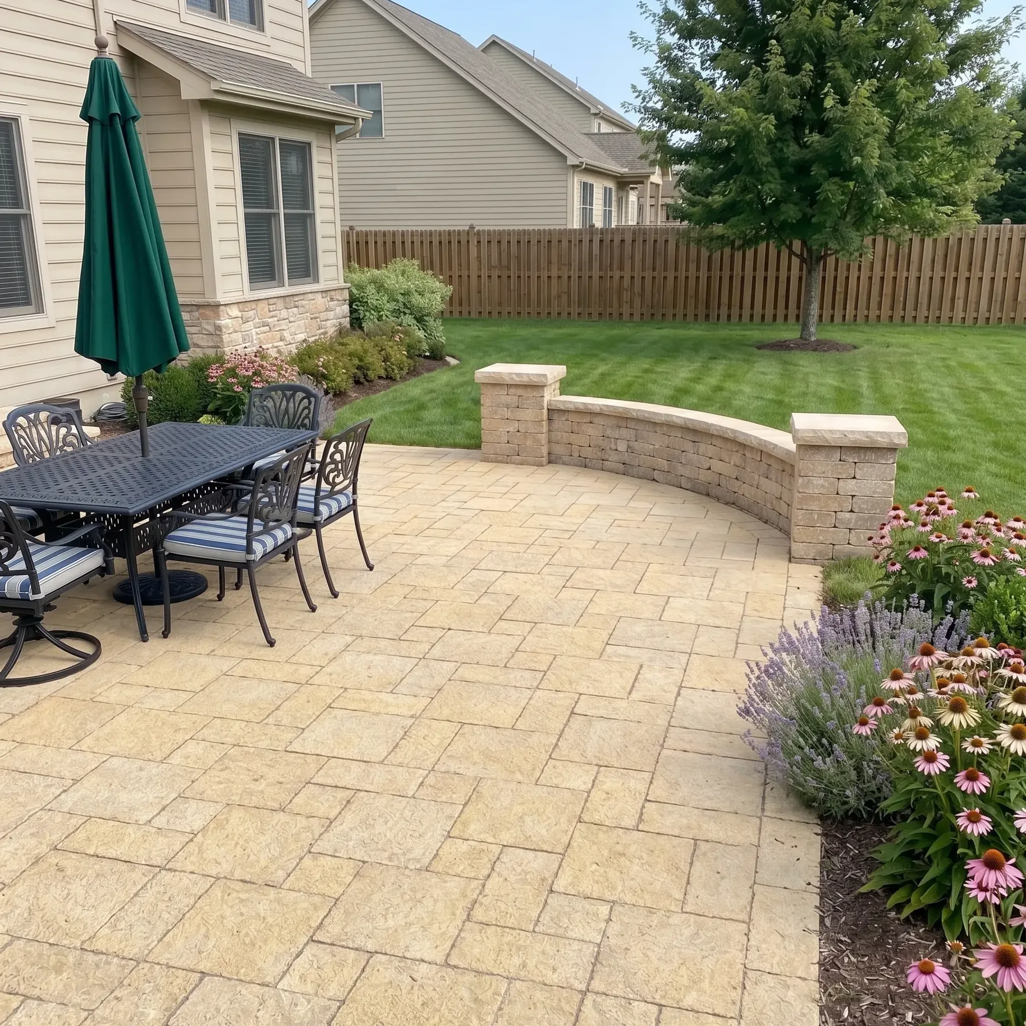 Golden tan textured ashlar-pattern pavers on a patio with curved stone seating wall, pillars, dining set, lawn, fence and…