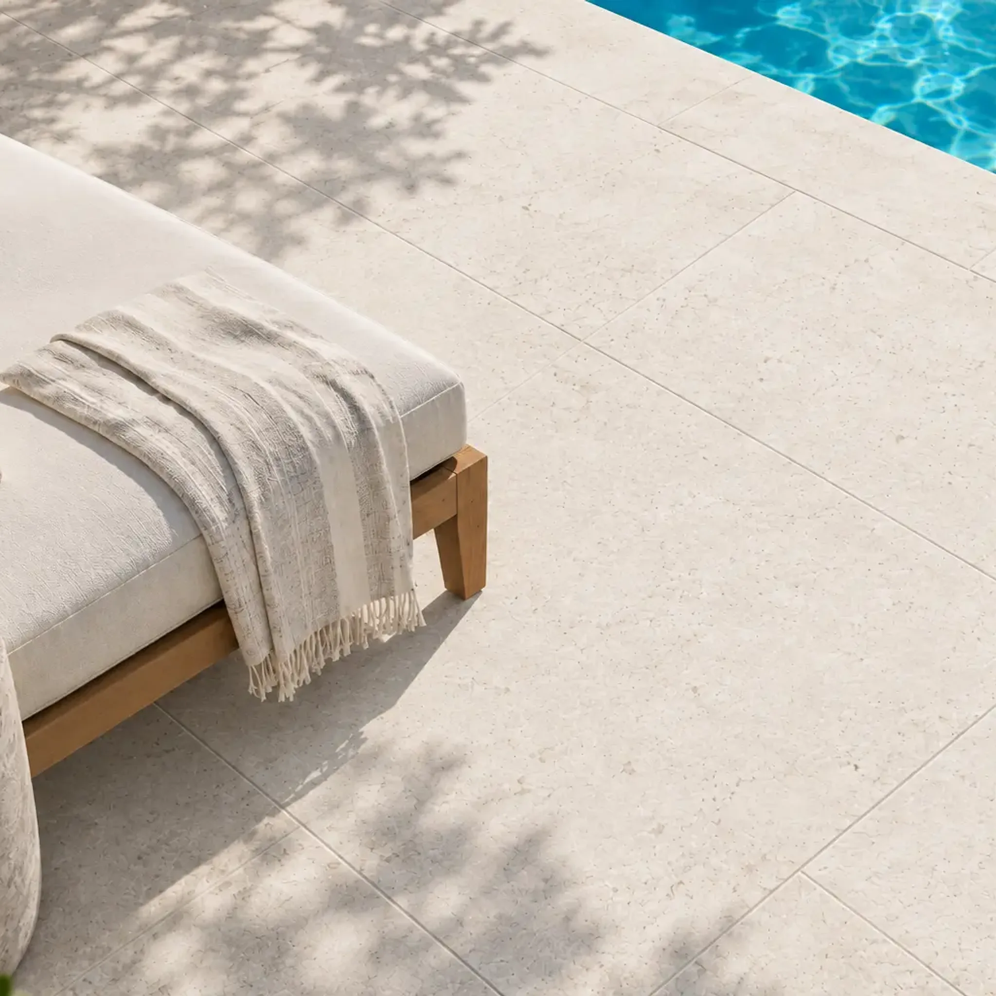 Off-white speckled limestone pavers beside a pool with a teak daybed, linen throw, stone side table and dappled tree shadows.