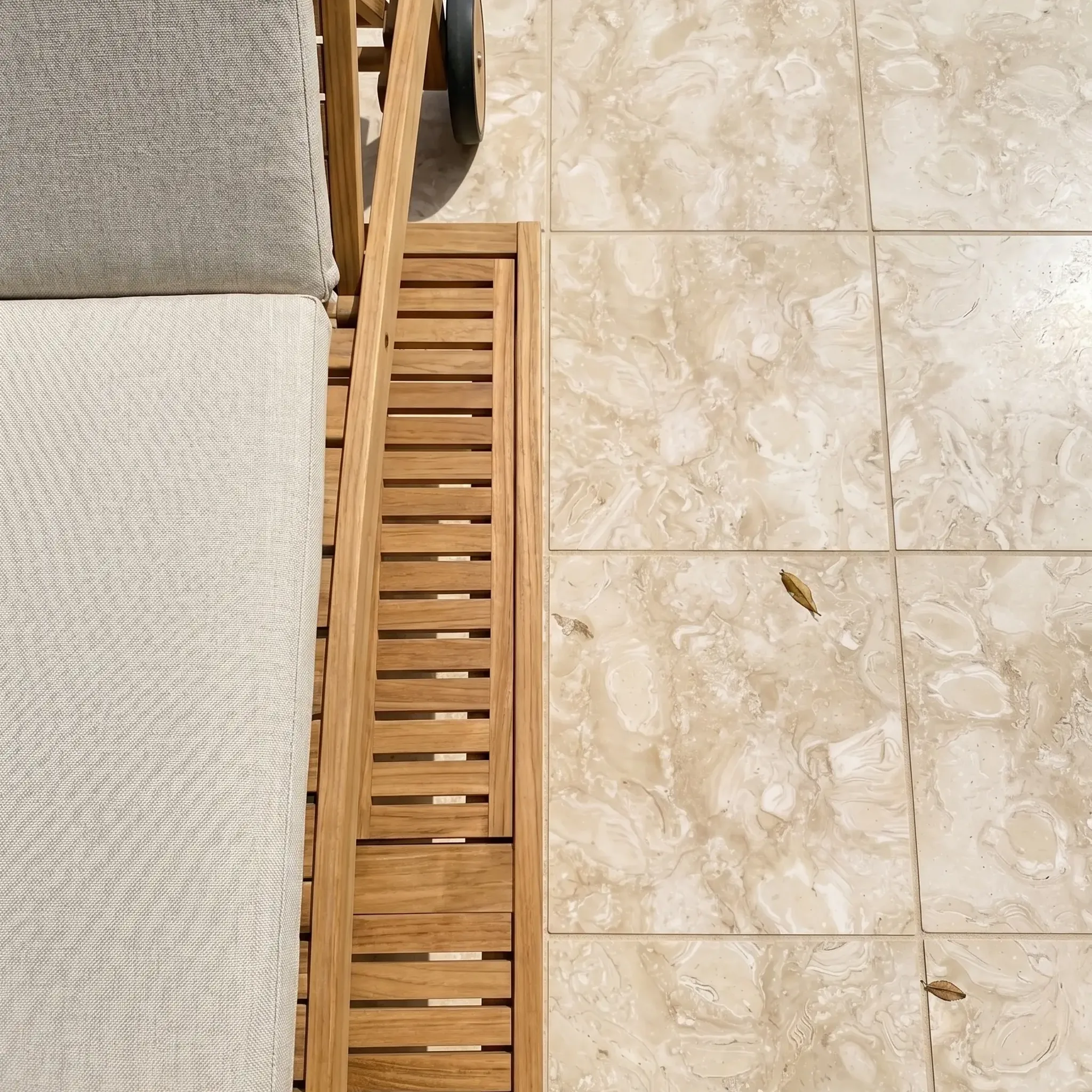 Overhead teak lounger with cushion and towel on beige fossil stone tiles with a few fallen leaves.