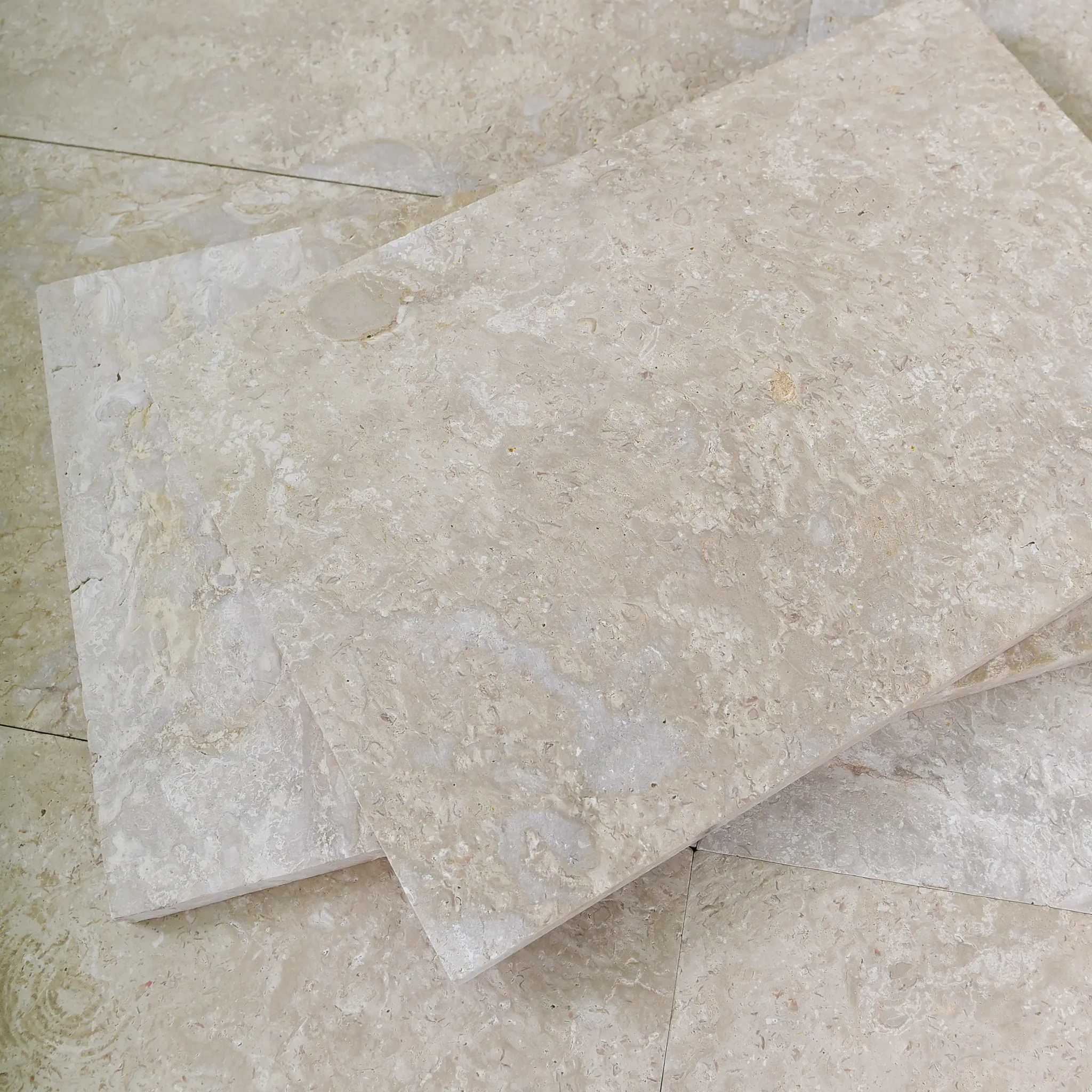 Two cream-beige brushed shellstone tiles stacked askew on a matching tiled floor below a wood-slat wall, showroom.