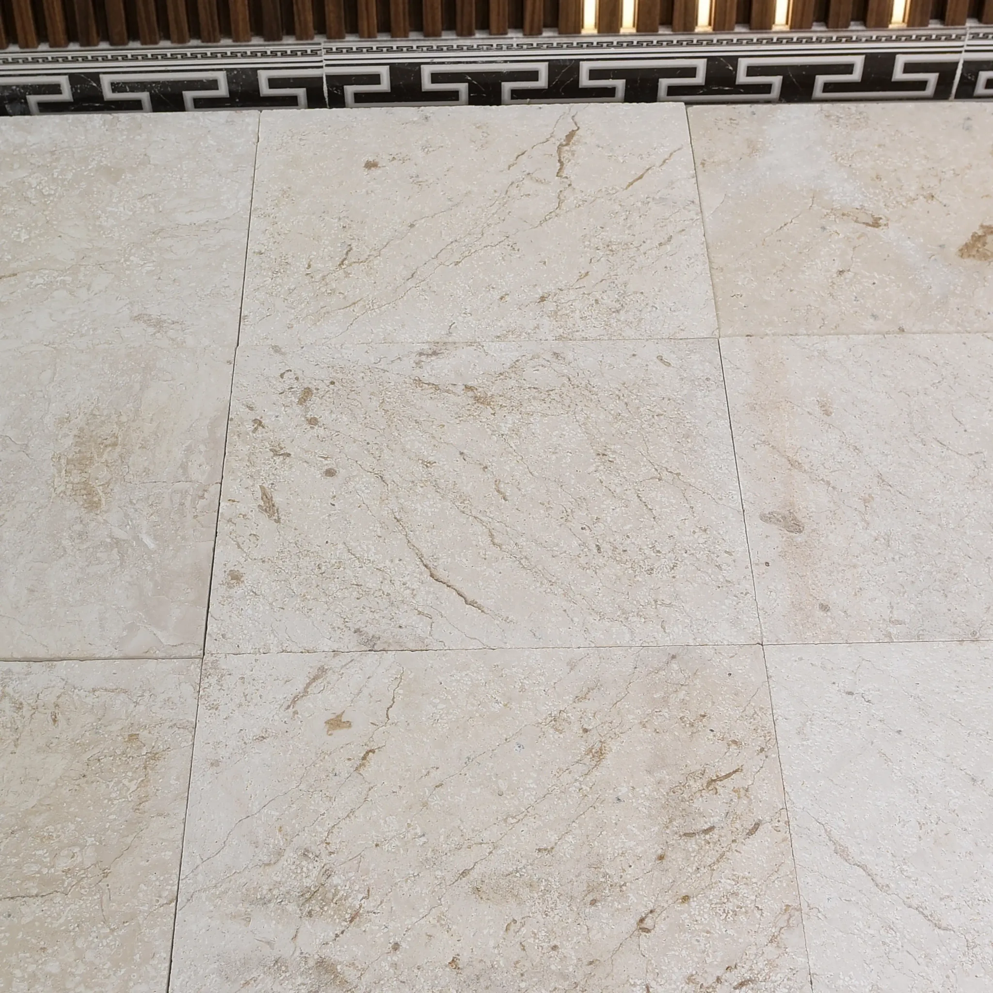 Cream limestone tiles with brown veining dry-laid below a Greek-key border and walnut slat wall with LED strips, white…