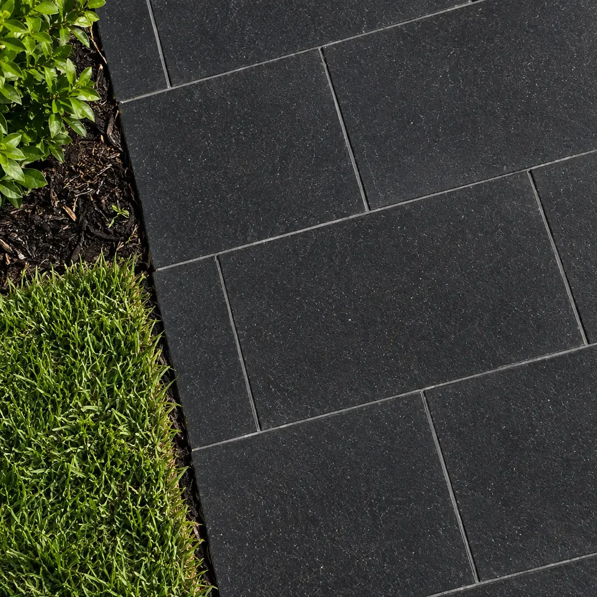 Top-down view of jet-black honed basalt pavers in running bond with thin joints beside lawn, mulch and a boxwood shrub.