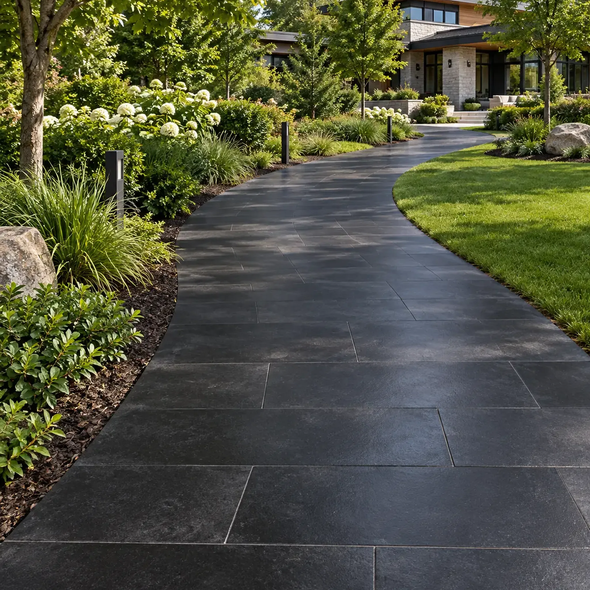 Curving black large-format stone slab walkway between planting beds and lawn toward a modern stone house, with boulders.