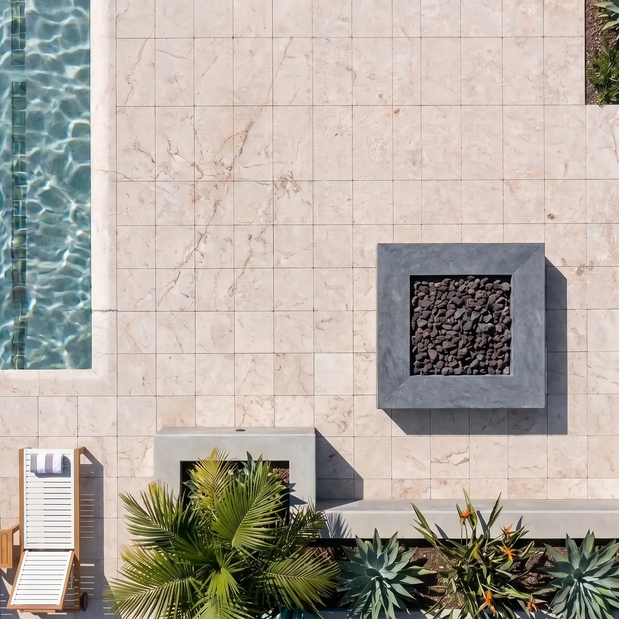 Top-down aerial of cream veined limestone pavers in a square grid beside a pool, three white loungers, fire pit and agaves.