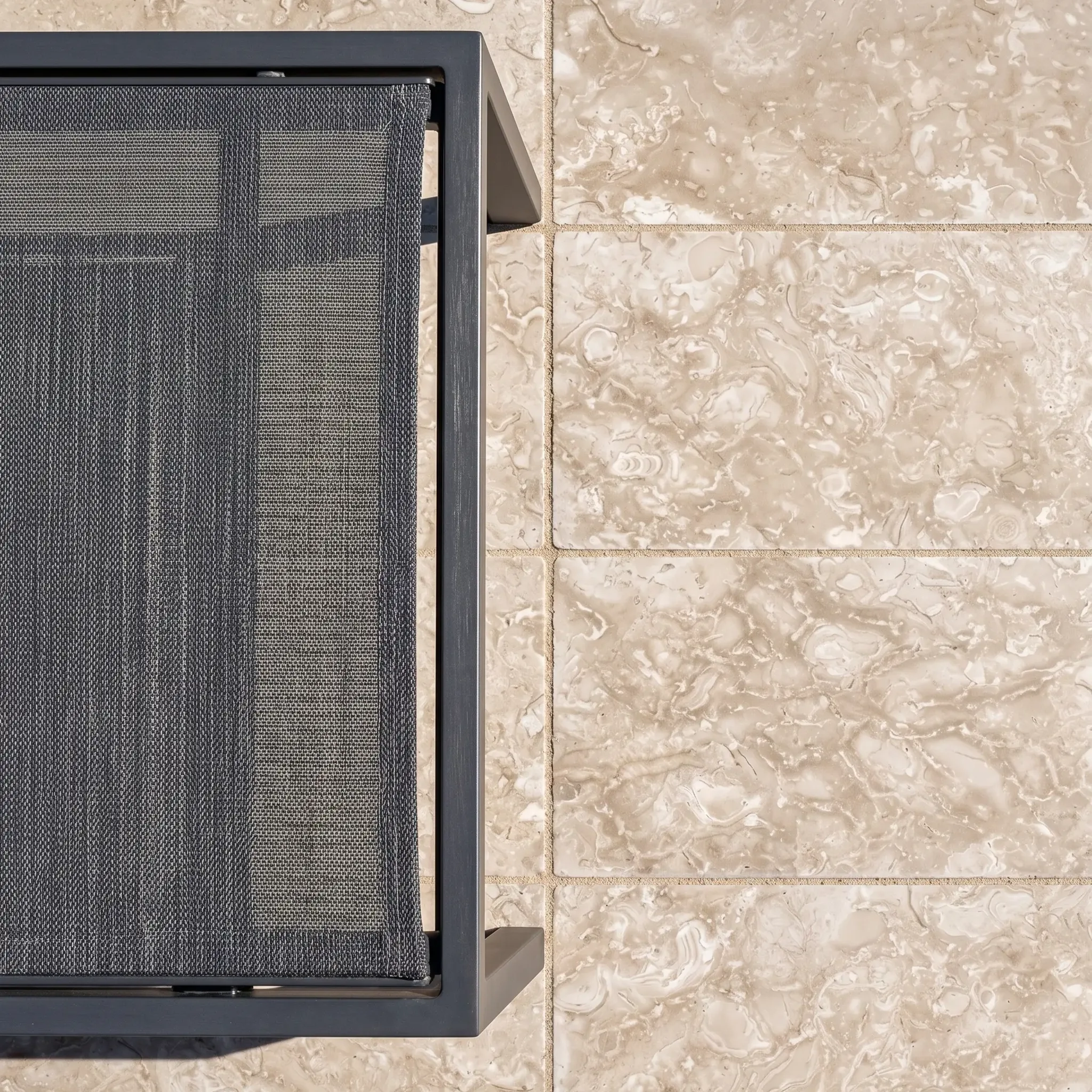 Macro overhead of a black sling lounger corner on beige fossil limestone pavers with sand-filled joints.