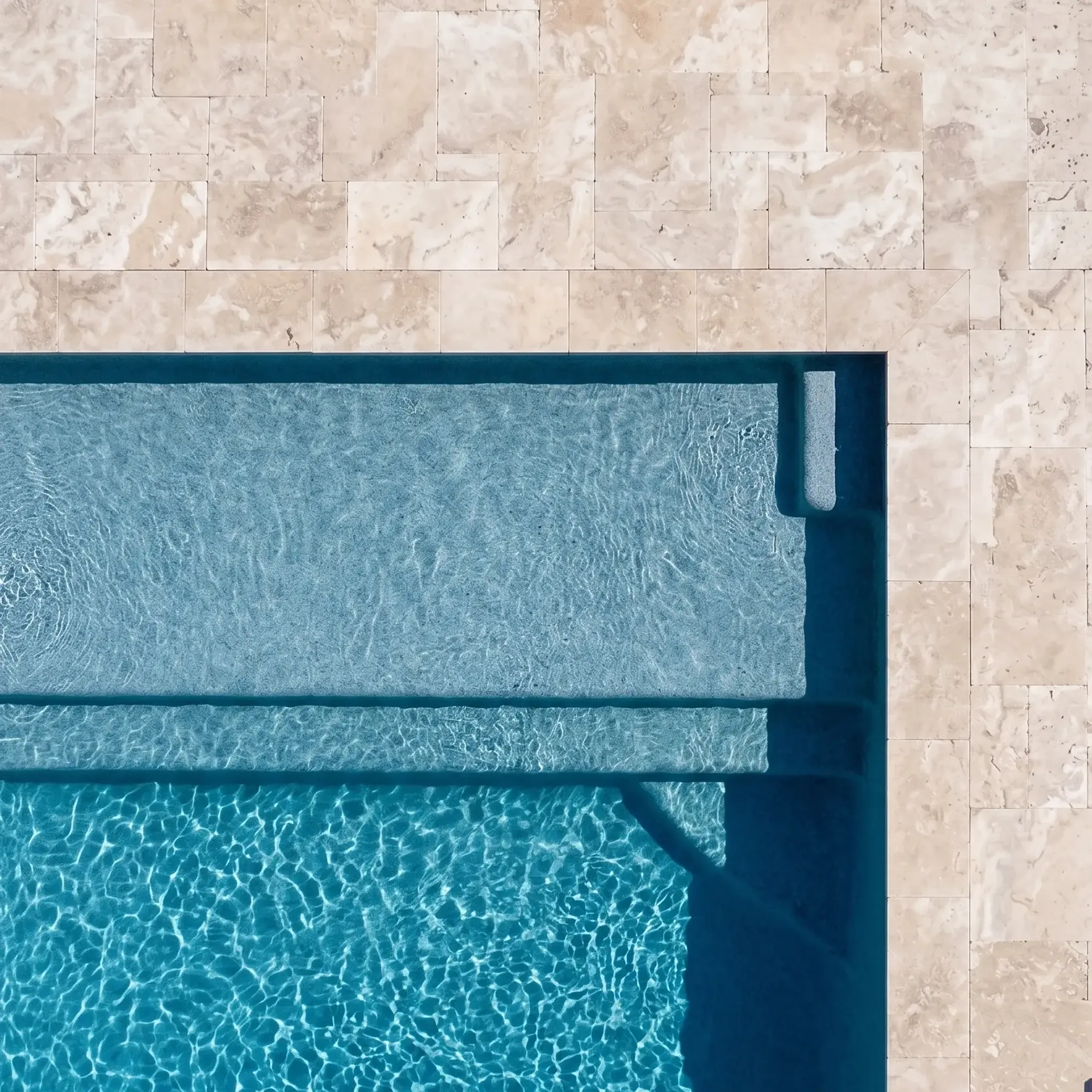 Aerial top-down of cream mottled stone pavers in French pattern around a pool corner with tanning ledge and steps.