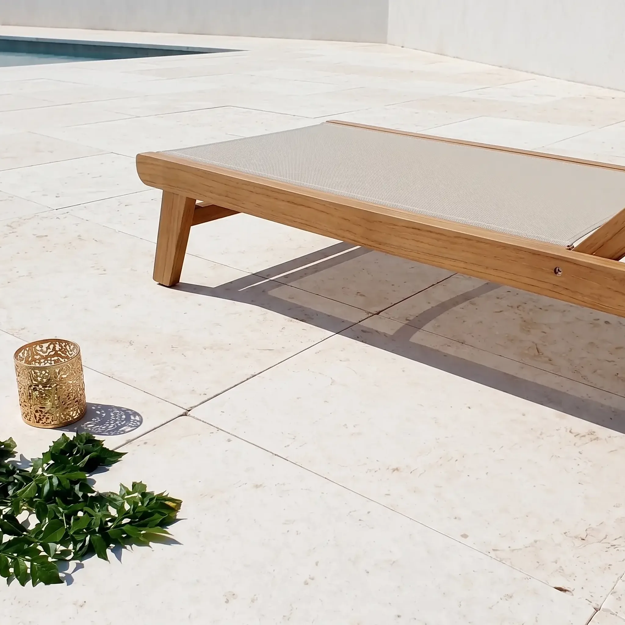 Bright photo of white limestone pavers with pitted surface, teak sling lounger, gold lantern and a leafy branch beside a…