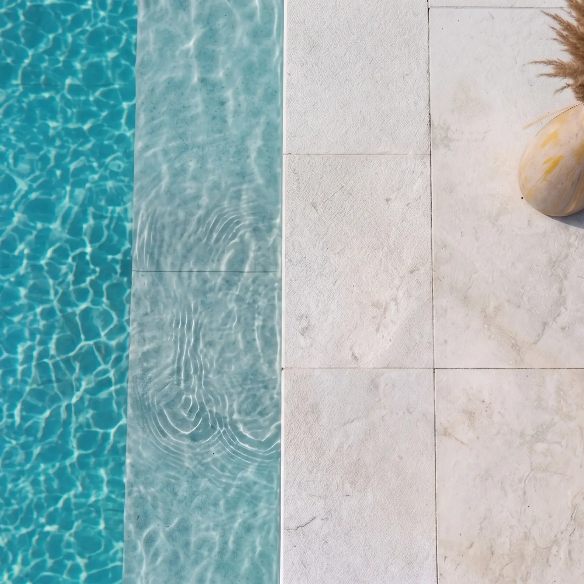 Top-down aerial of off-white limestone pavers at a pool edge with a submerged sun ledge and a pampas vase casting a shadow.