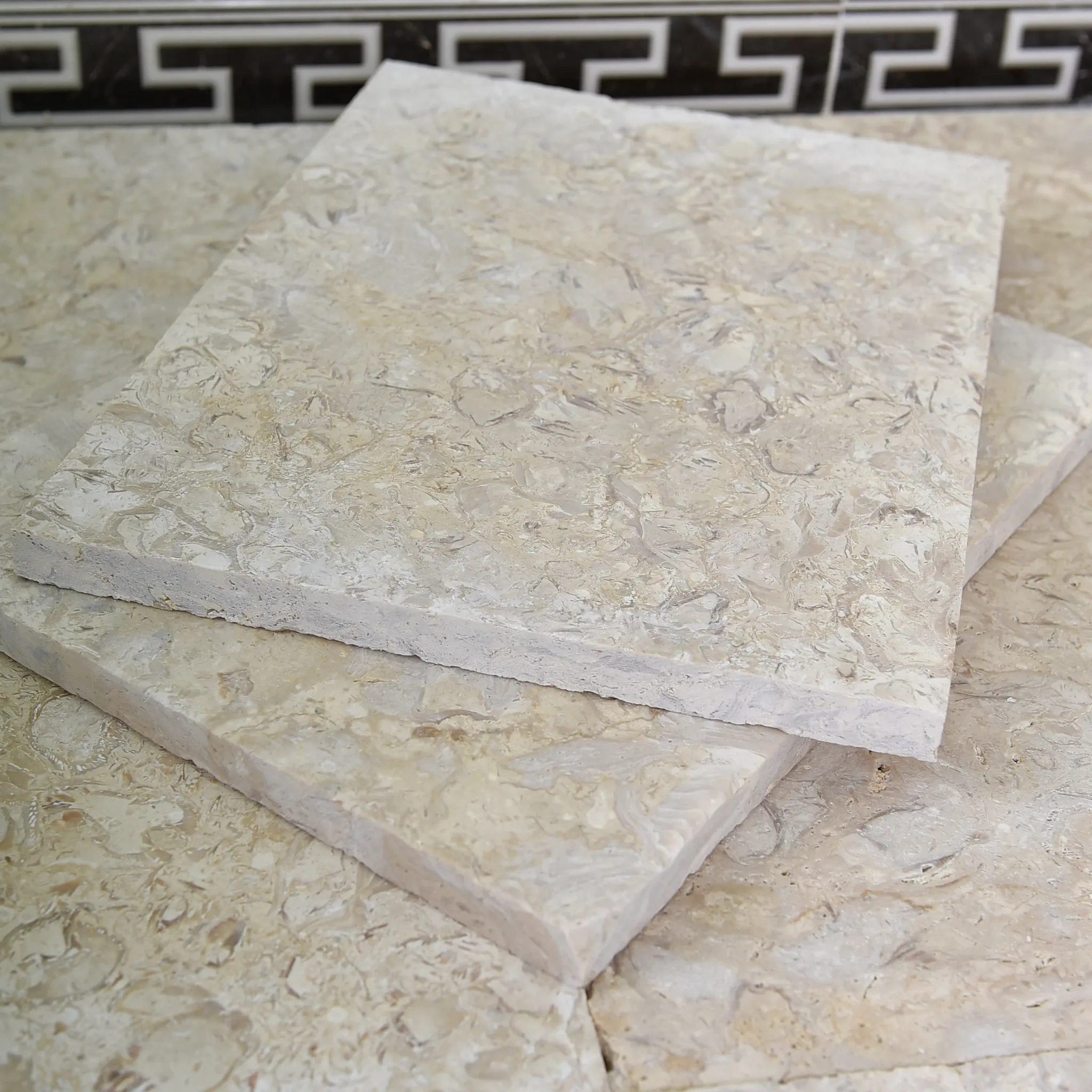 Two brushed fossil beige limestone tiles stacked on a matching dry-laid floor before Greek-key skirting and wood slat wall.