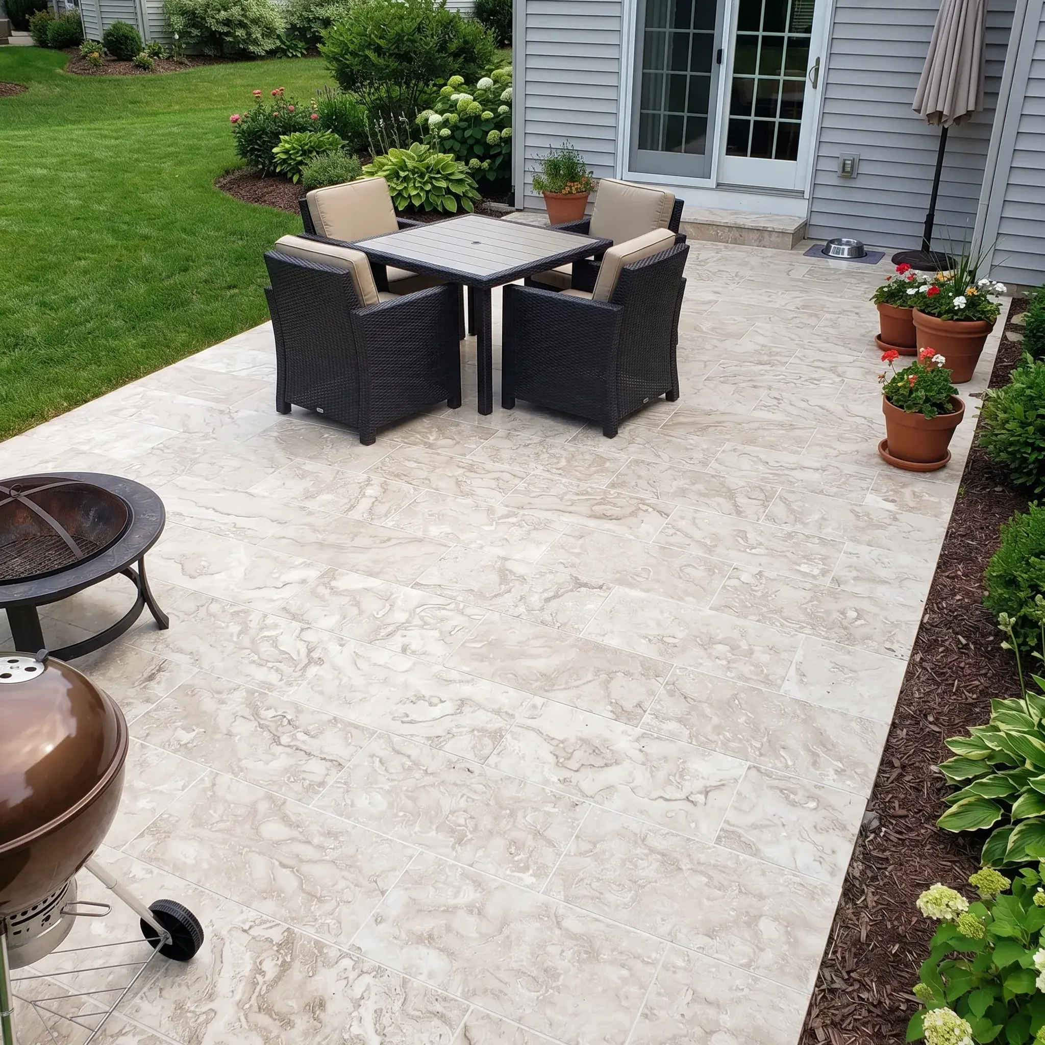 Cream swirl-veined limestone tile patio with wicker dining set, grill, fire pit and potted geraniums by a grey house.