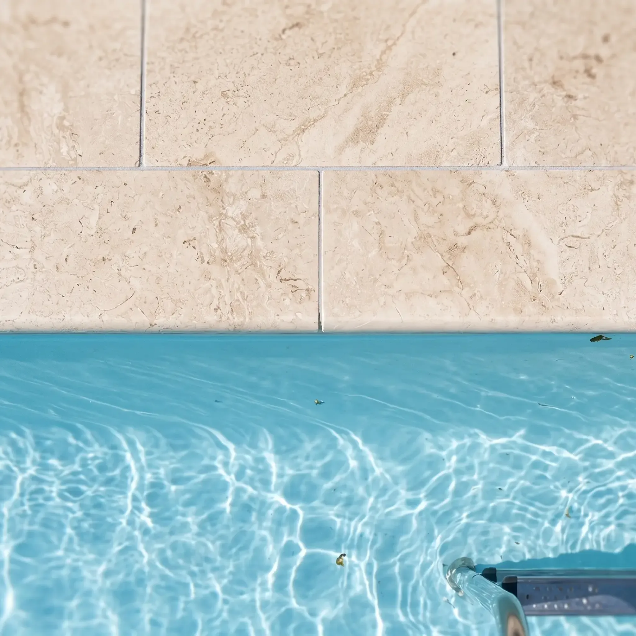Close-up of cream limestone pool coping and pavers with a bullnose edge over turquoise water, pool ladder rail at lower…
