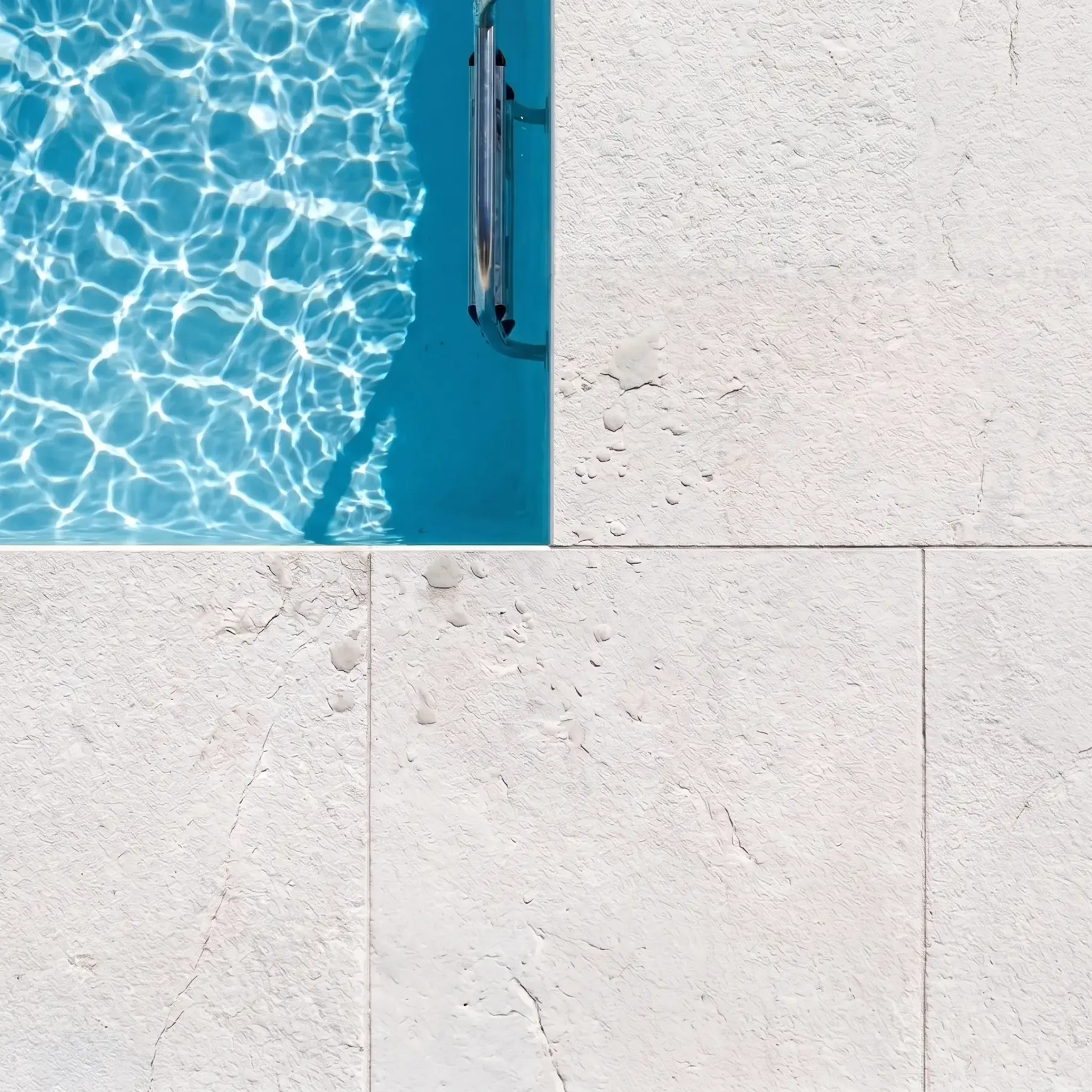 Overhead view of white limestone pavers with pitted texture meeting a pool edge with chrome ladder, bright sunlight and…