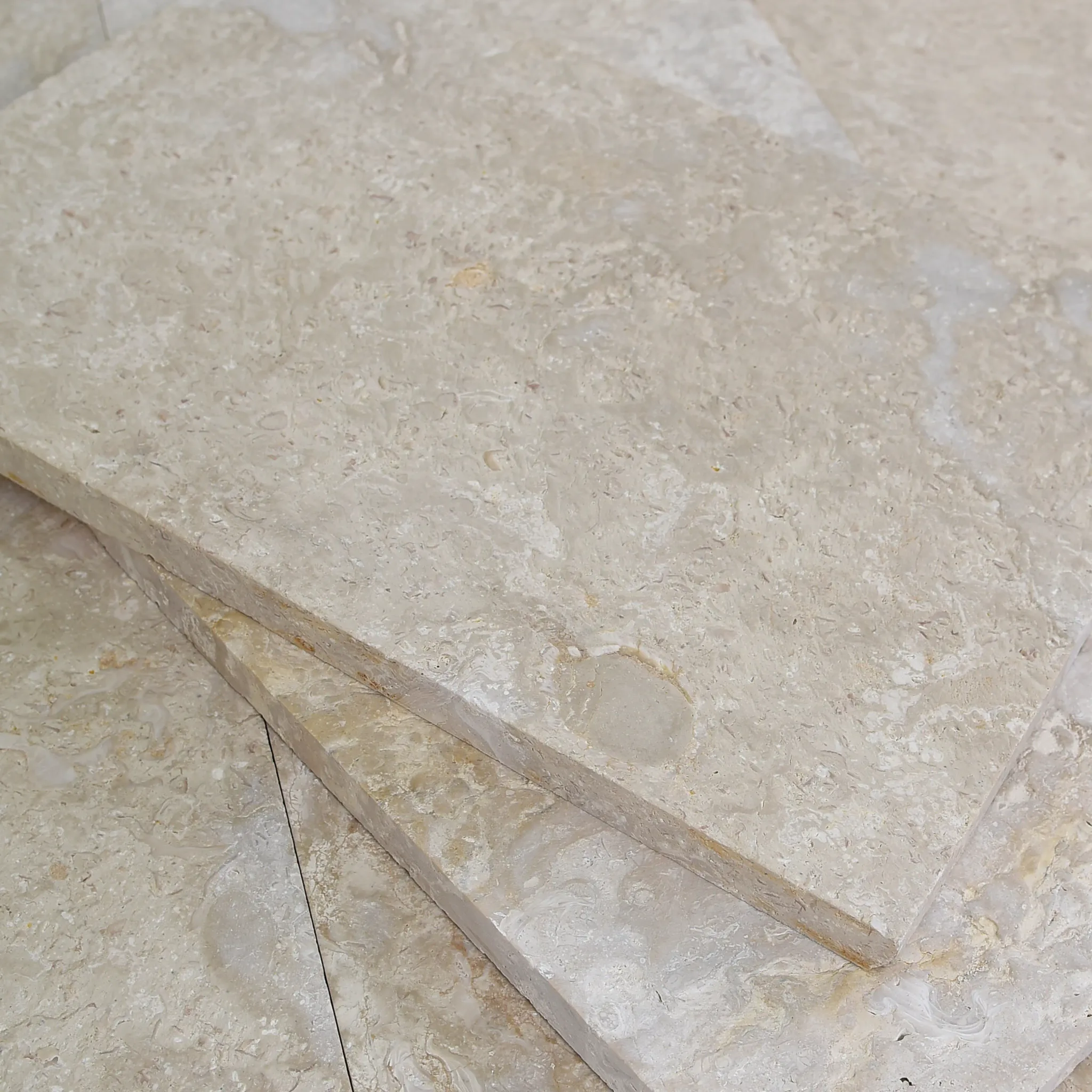 Stacked beige fossil shellstone pavers with sawn edges showing thickness on a tiled studio floor by a wood slat wall.